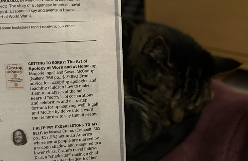 My cat Novella, sleeping next to a print copy of the NYT announcing the publication of Getting to Sorry in paperback